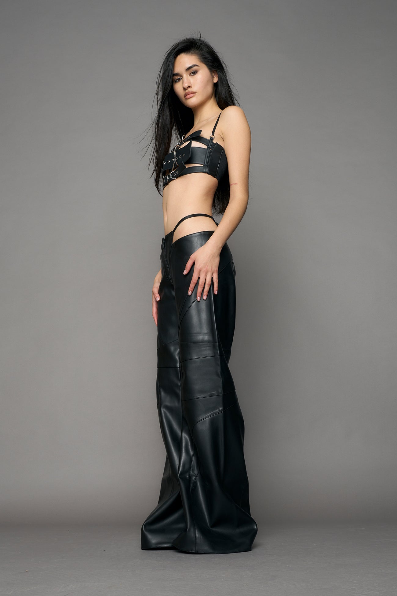Low-rise flared pants in black faux leather with bold side cut-outs and thong strap detailing. Featuring moto-style paneling, front zip closure, and a sculpted fit. A statement piece for rave, nightlife, and runway-inspired looks.