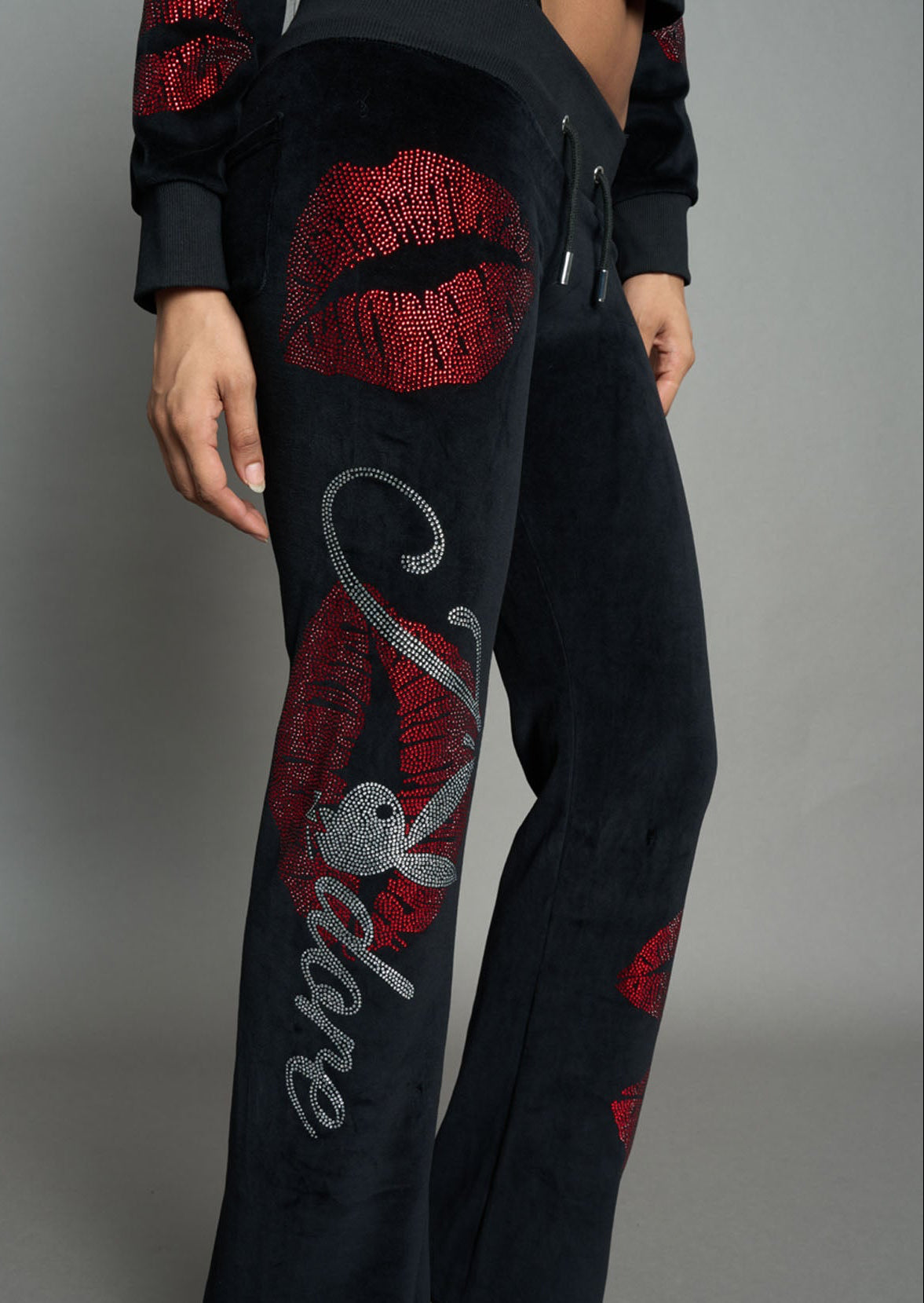 model wearing black velvet tracksuit pants with rhinestone embroidery of red lip prints and the lettering "J'adore Playboy" ( "a" is a Playboy Bunny Head", standing against a solid gray studio background.