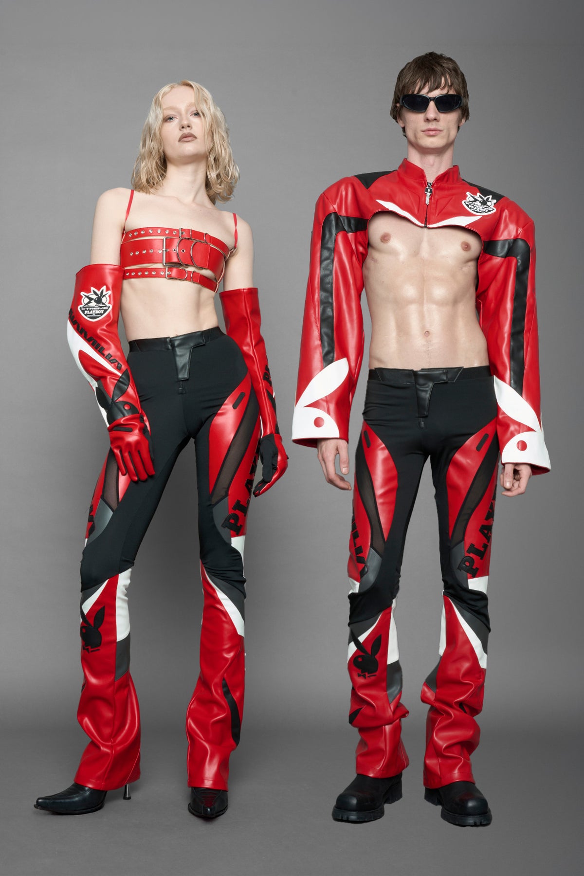 model wearing a red and black form fitted Pants with PU Leather Embroidery of 'Namilia', 'Playboy'. The pants have black Mesh fabric and dark grey PU leather panels and are slightly flared from knee to hem
