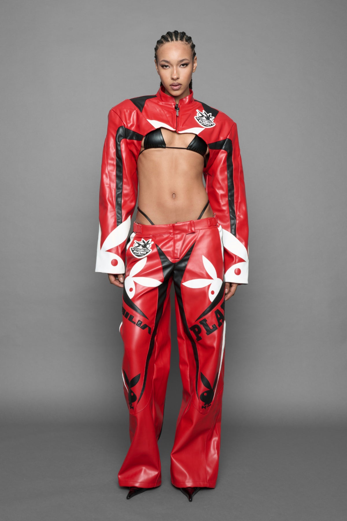 model wearing a vegan leather cropped jacket with broad shoulders in red with black and white paneling. The jacket has PU leather embroidery of 'Namilia' and 'Playboy' with a key formed zipper pull and a rubbertag on the left breast.