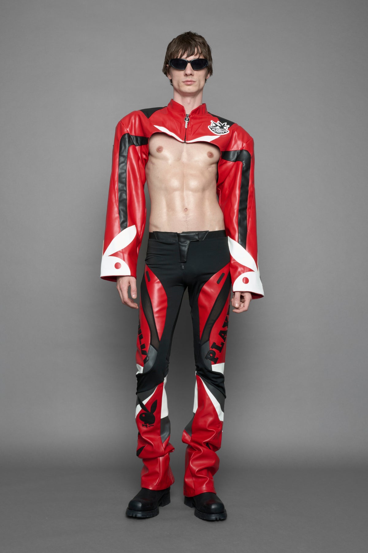 model wearing a red and black form fitted Pants with PU Leather Embroidery of 'Namilia', 'Playboy'. The pants have black Mesh fabric and dark grey PU leather panels and are slightly flared from knee to hem