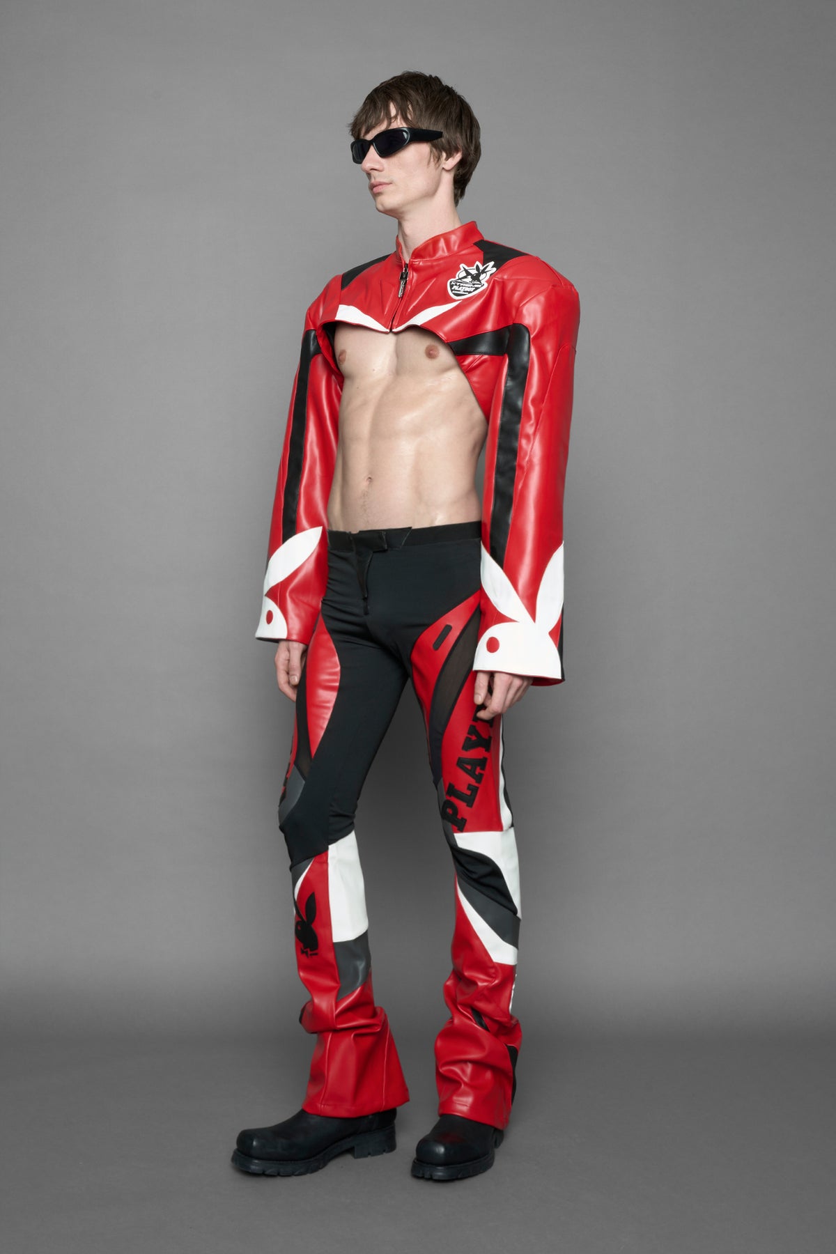 model wearing a red and black form fitted Pants with PU Leather Embroidery of 'Namilia', 'Playboy'. The pants have black Mesh fabric and dark grey PU leather panels and are slightly flared from knee to hem