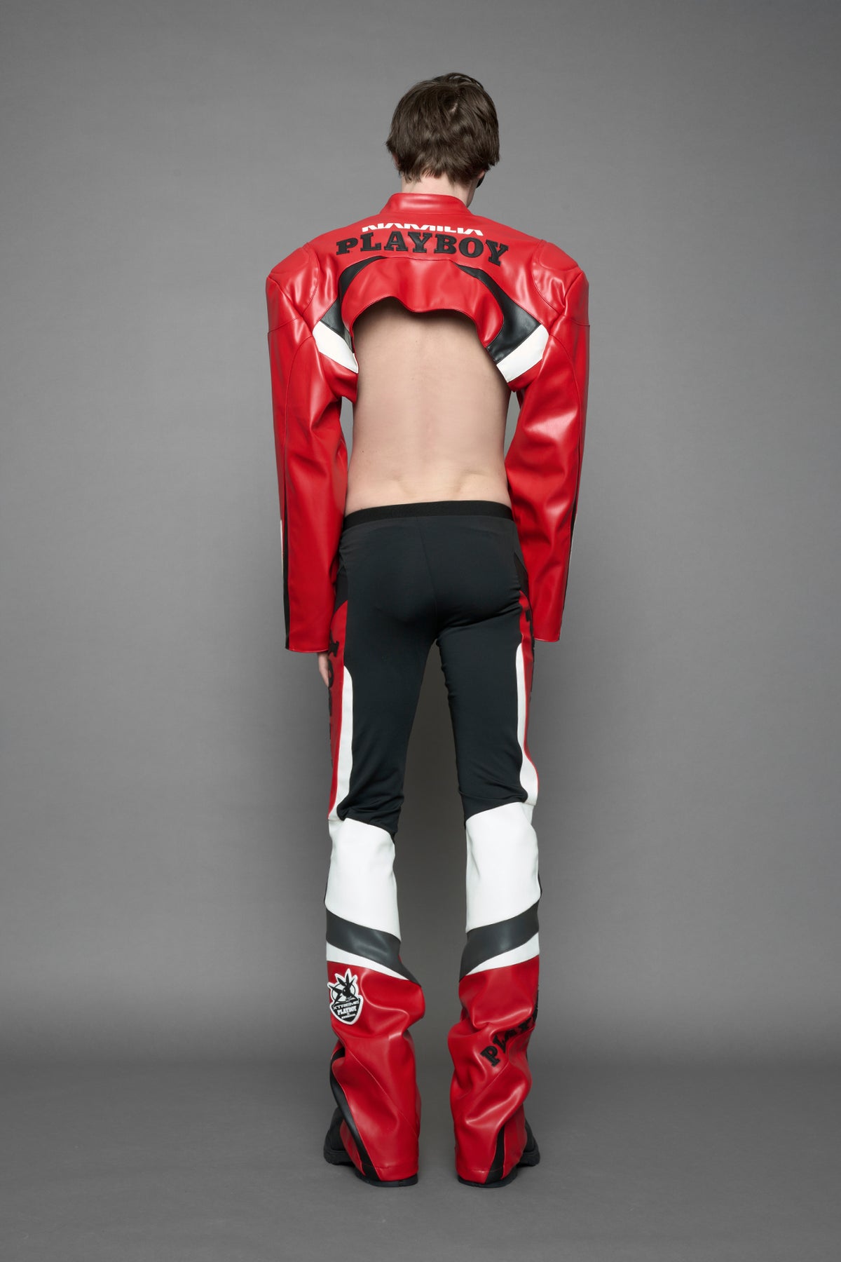 model wearing a red and black form fitted Pants with PU Leather Embroidery of 'Namilia', 'Playboy'. The pants have black Mesh fabric and dark grey PU leather panels and are slightly flared from knee to hem