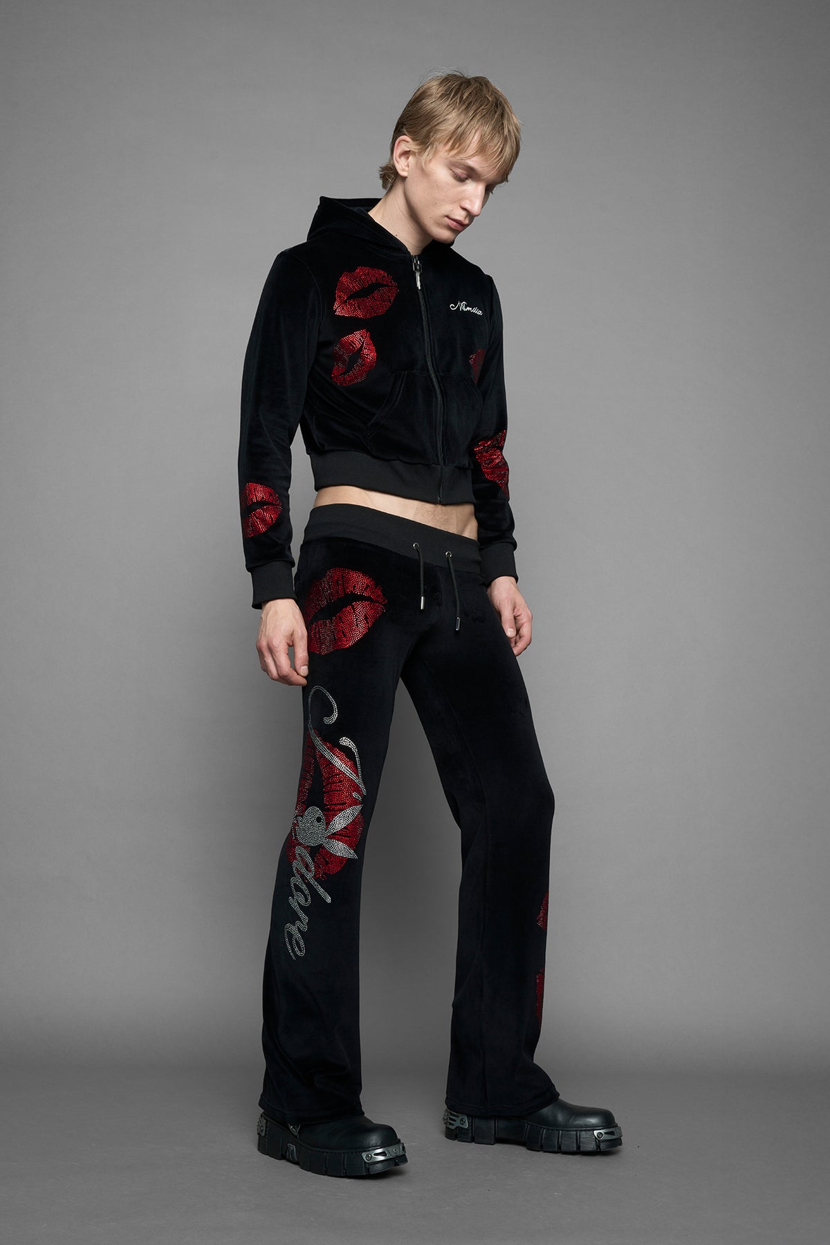 A male model wearing black velvet tracksuit pants with rhinestone embroidery of red lip prints and the lettering "J'adore Playboy" ( "a" is a Playboy Bunny Head", standing against a solid gray studio background.