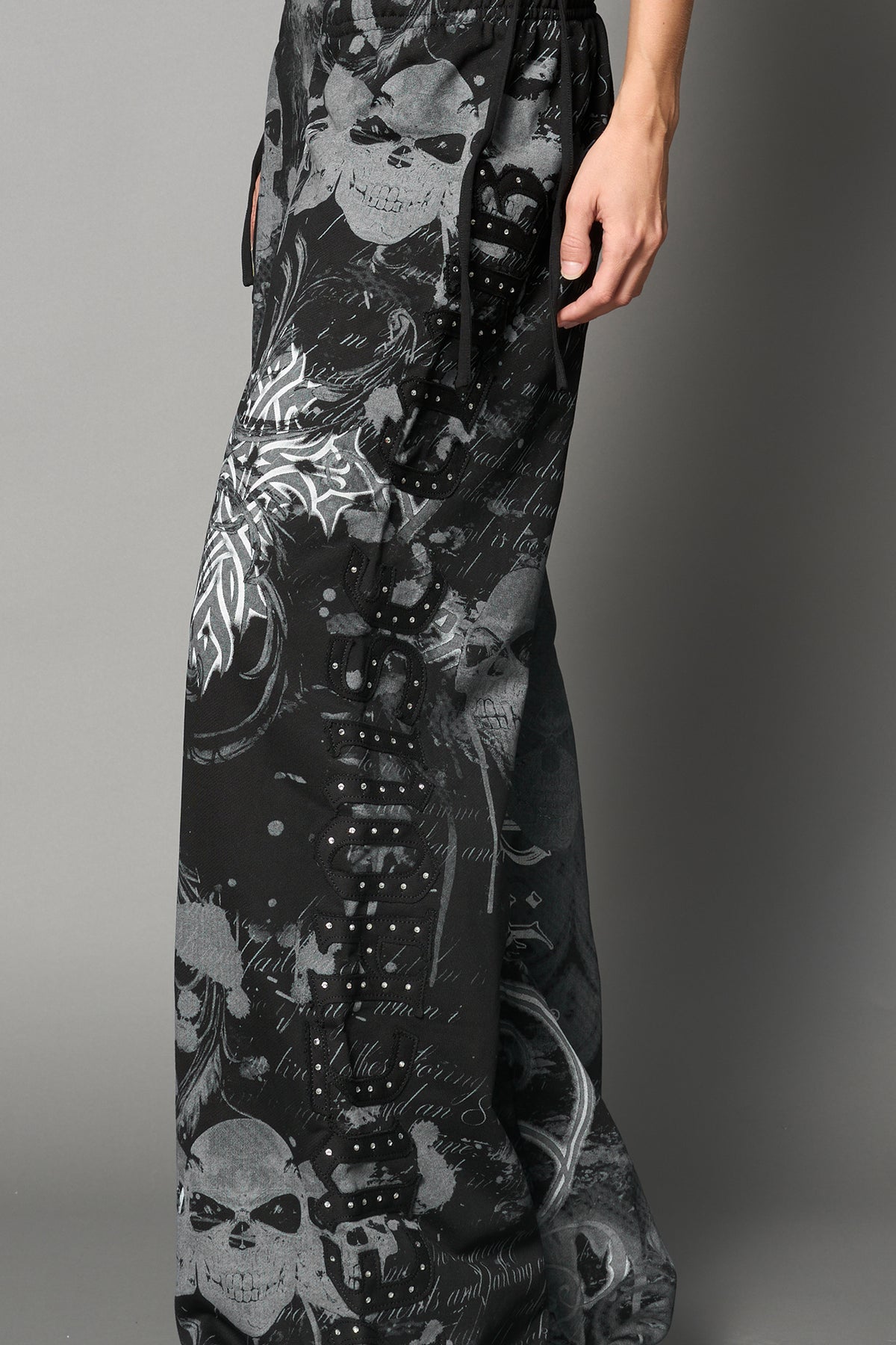 Model wearing low-rise black print wide-leg pants featuring a V-shaped waistband and detachable thong strap detail.