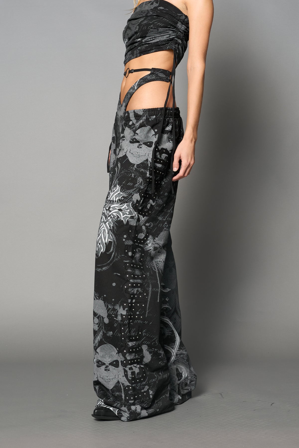 Model wearing low-rise black print wide-leg pants featuring a V-shaped waistband and detachable thong strap detail.