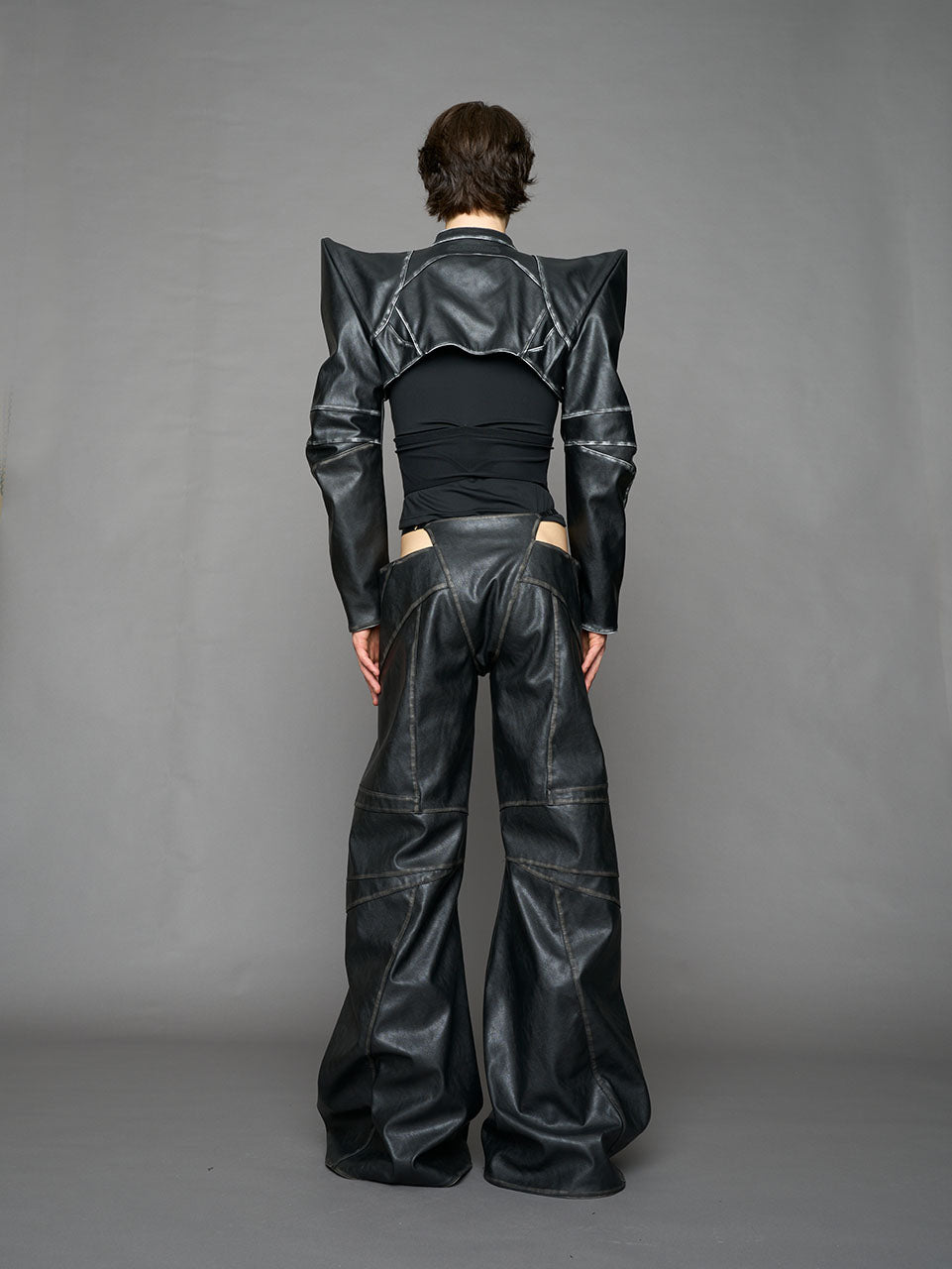 Model wearing a black vegan leather spike shoulder bolero jacket with cut-out design on a gray background