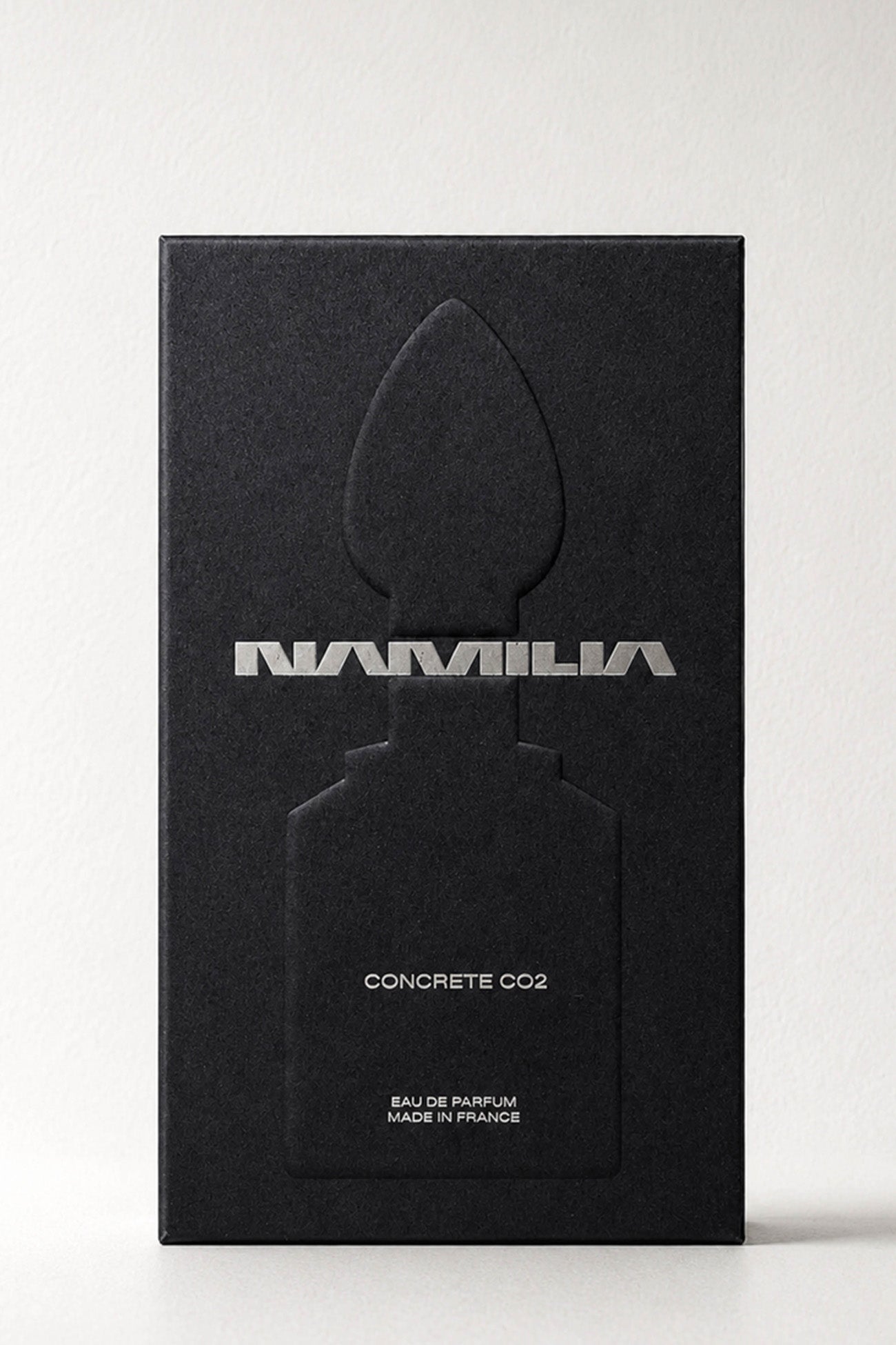 A bottle of Namila Concrete CO2 Eau de Parfum with a reflective silver plug-shaped cap and a clear glass base showing a pale yellow liquid, set against a solid black background.