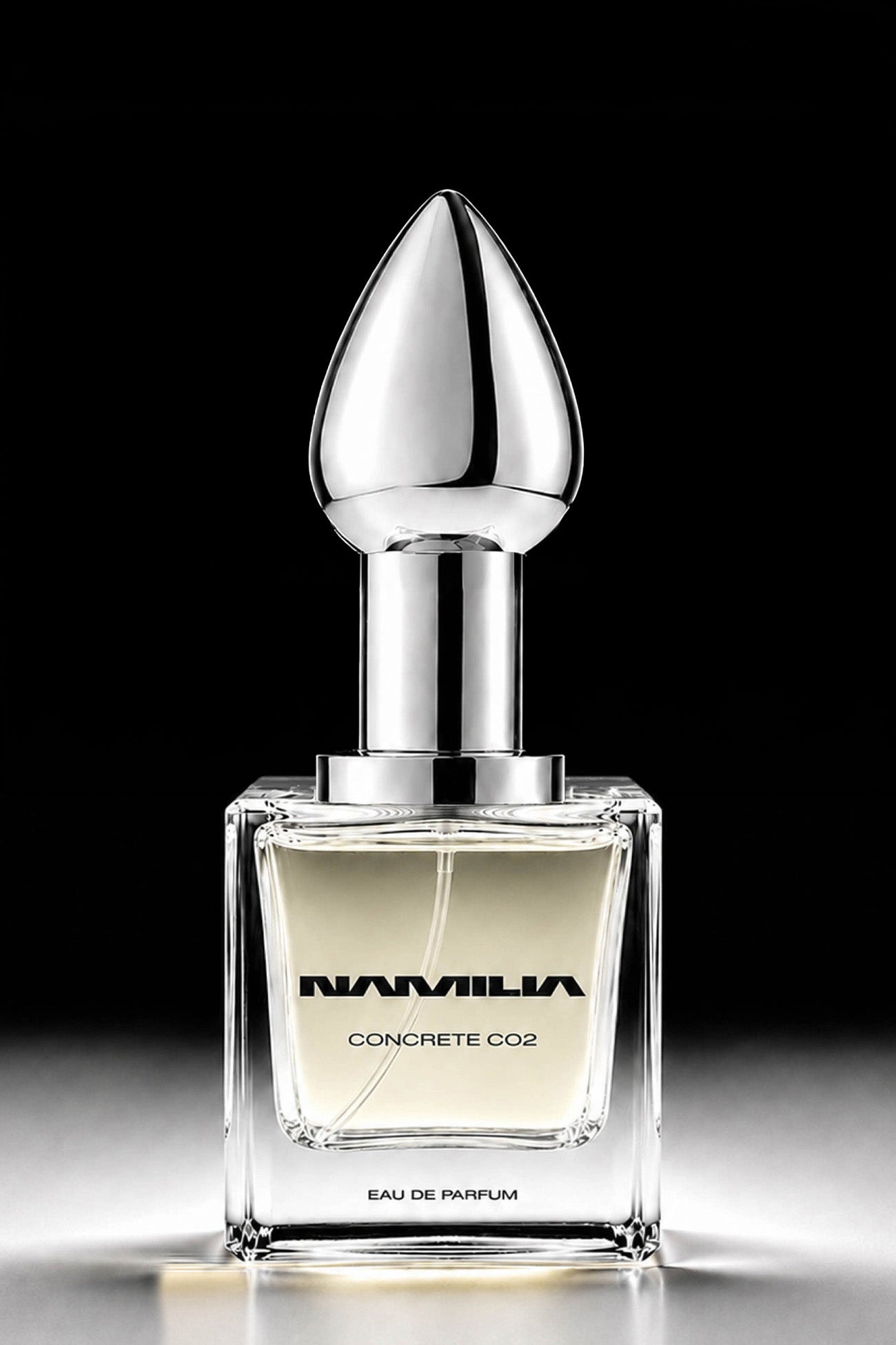 A bottle of Namila Concrete CO2 Eau de Parfum with a reflective silver plug-shaped cap and a clear glass base showing a pale yellow liquid, set against a solid black background.