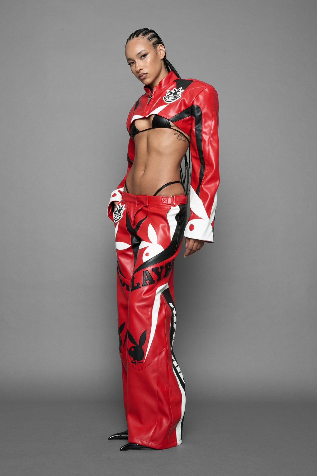 model wearing a vegan leather cropped jacket with broad shoulders in red with black and white paneling. The jacket has PU leather embroidery of 'Namilia' and 'Playboy' with a key formed zipper pull and a rubbertag on the left breast.