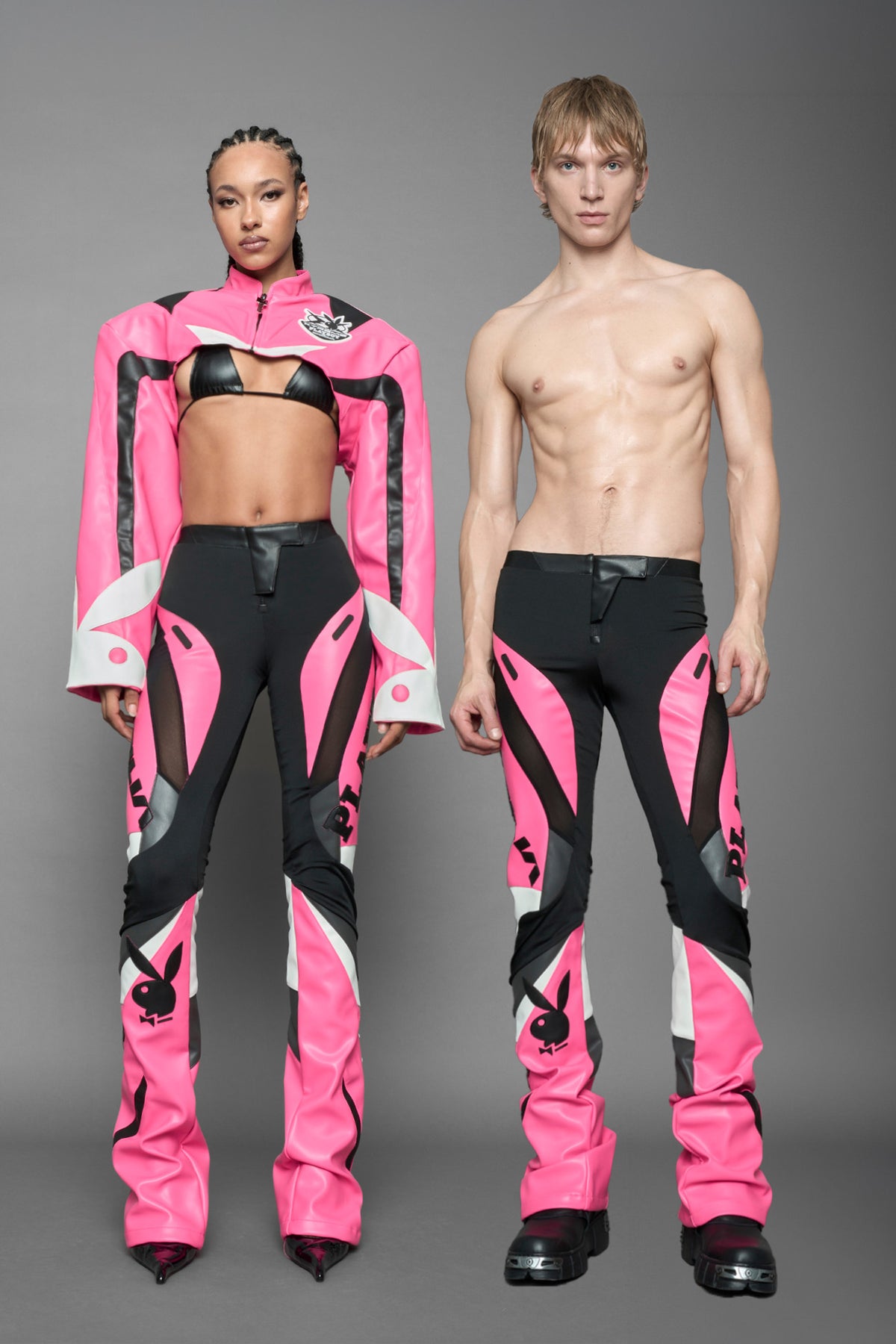 model wearing a pink and black form fitted Pants with PU Leather Embroidery of 'Namilia', 'Playboy'. The pants have black Mesh fabric and dark grey PU leather  panels and are slightly flared from knee to hem.
