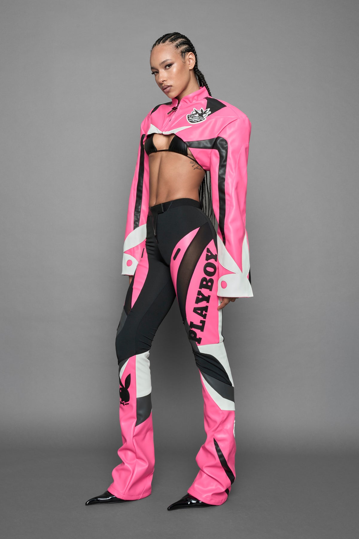 A female model wearing a pink and black form fitted Pants with PU Leather Embroidery of 'Namilia', 'Playboy'. The pants have black Mesh fabric and dark grey PU leather  panels and are slightly flared from knee to hem.