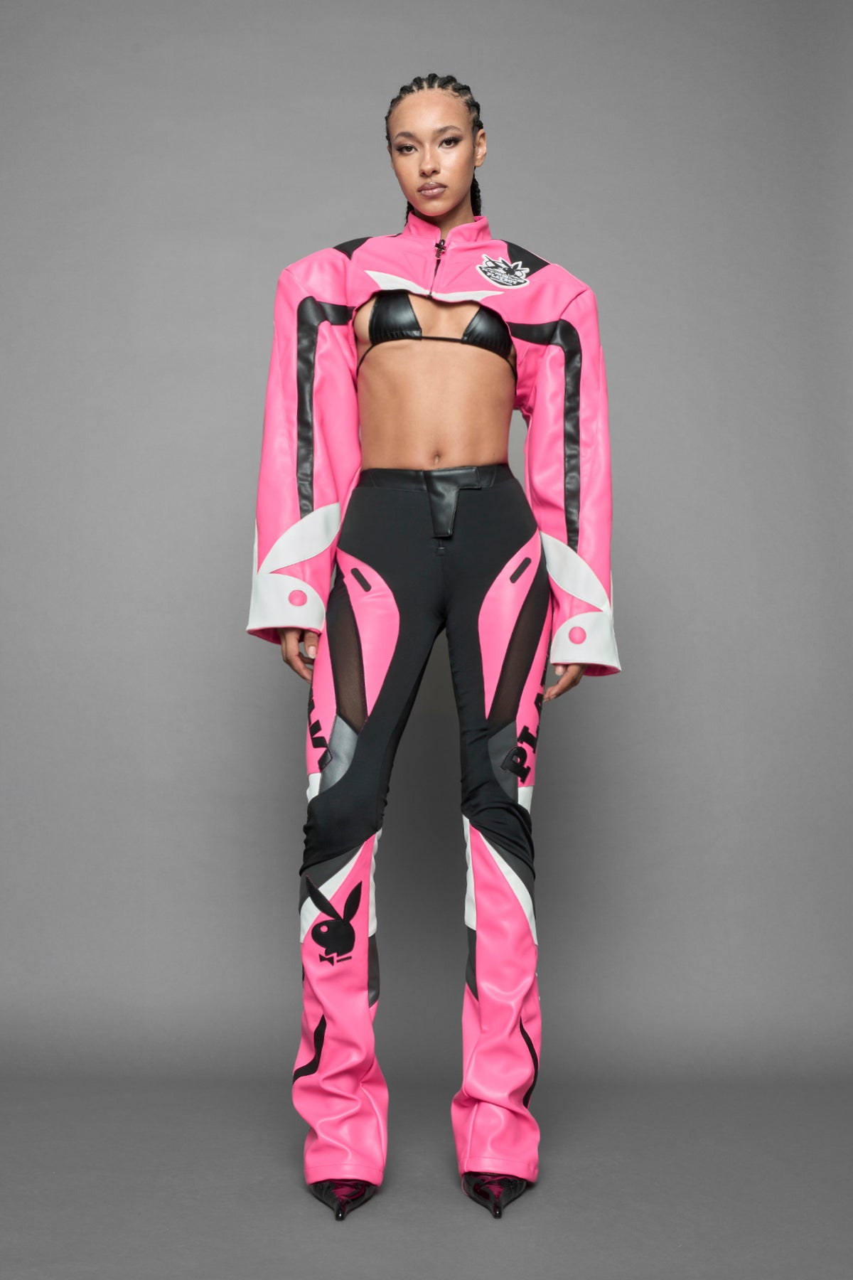 A female model wearing a pink and black form fitted Pants with PU Leather Embroidery of 'Namilia', 'Playboy'. The pants have black Mesh fabric and dark grey PU leather  panels and are slightly flared from knee to hem.